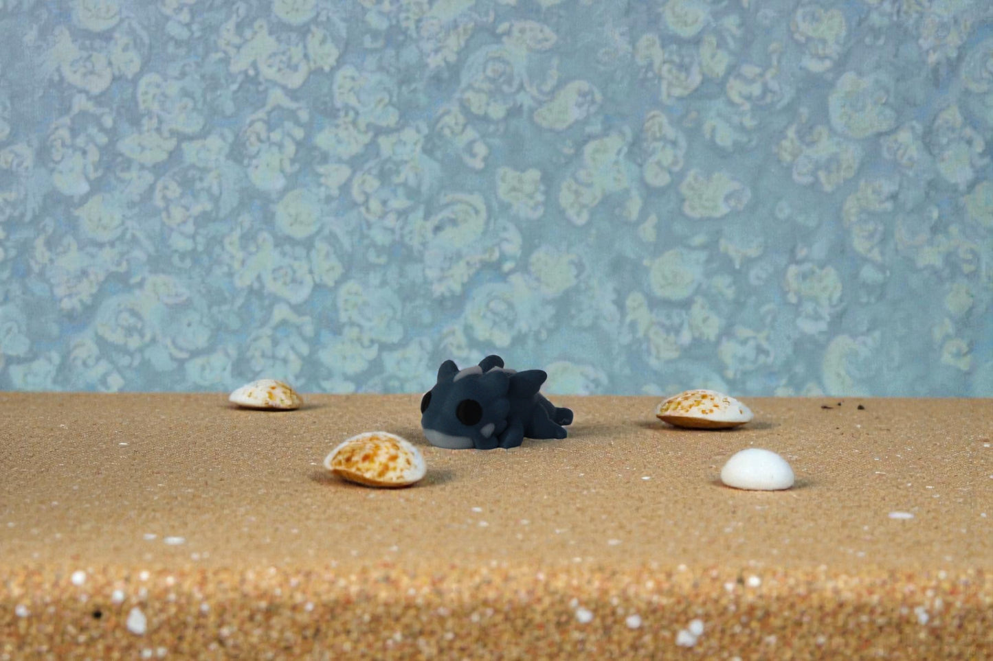 Mini articulated water dragon collectible in matte denim blue PLA, 3D printed in small batches, shown resting on sandy surface with seashell accents, affordable fantasy desk and shelf decor for collectors and themed collections.
