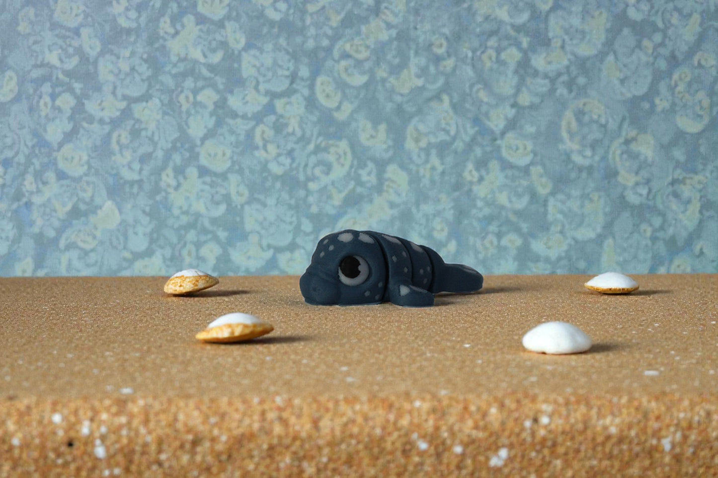 Front angled view of mini articulated water dragon collectible in matte denim blue PLA, 3D printed in small batches, displayed on sandy surface with seashell accents, affordable fantasy decor piece for shelf styling and dragon collections.
