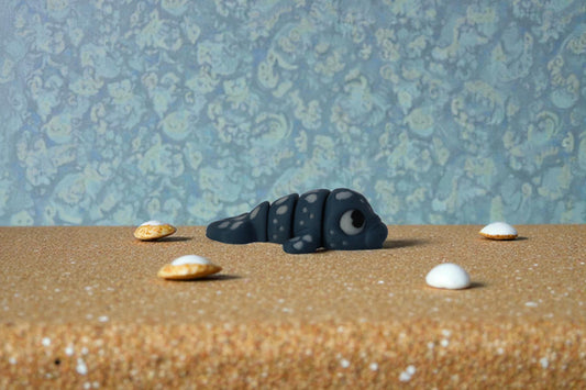 Mini articulated water dragon collectible in matte denim blue PLA, 3D printed in small batches, shown in side profile on sandy surface with seashell accents, affordable fantasy shelf decor for collectors and themed collections.
