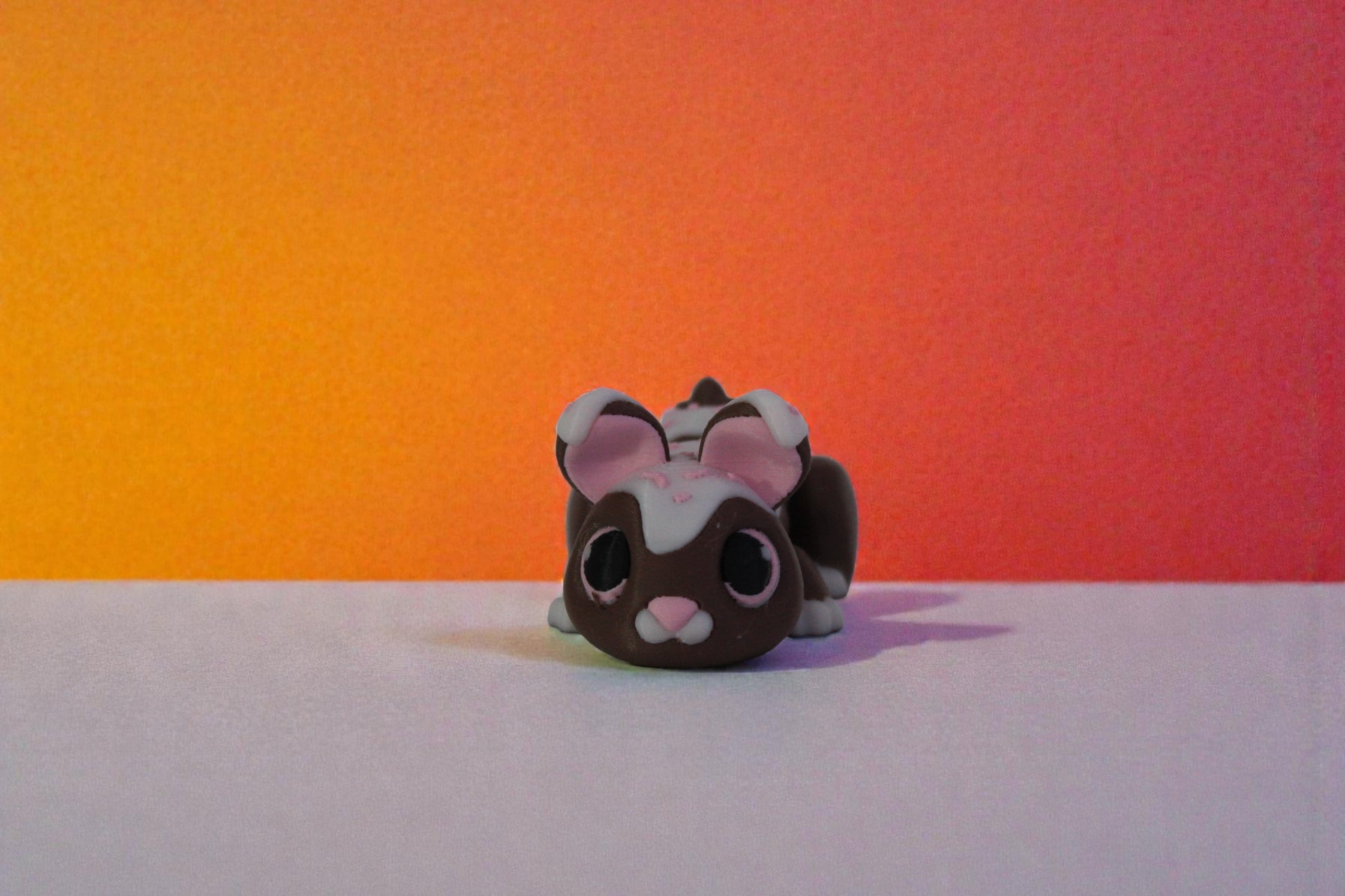 Mini articulated chocolate bunny decor with white glaze and pink sprinkles, front seated view on neutral surface with warm gradient background, 3D printed collectible.